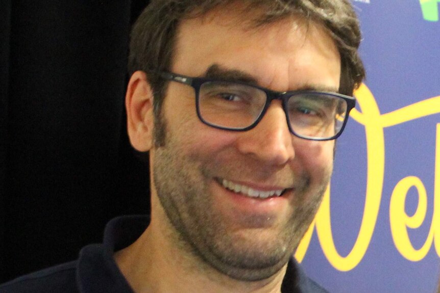 A man with dark hair and dark eyeglasses smiles at the camera.