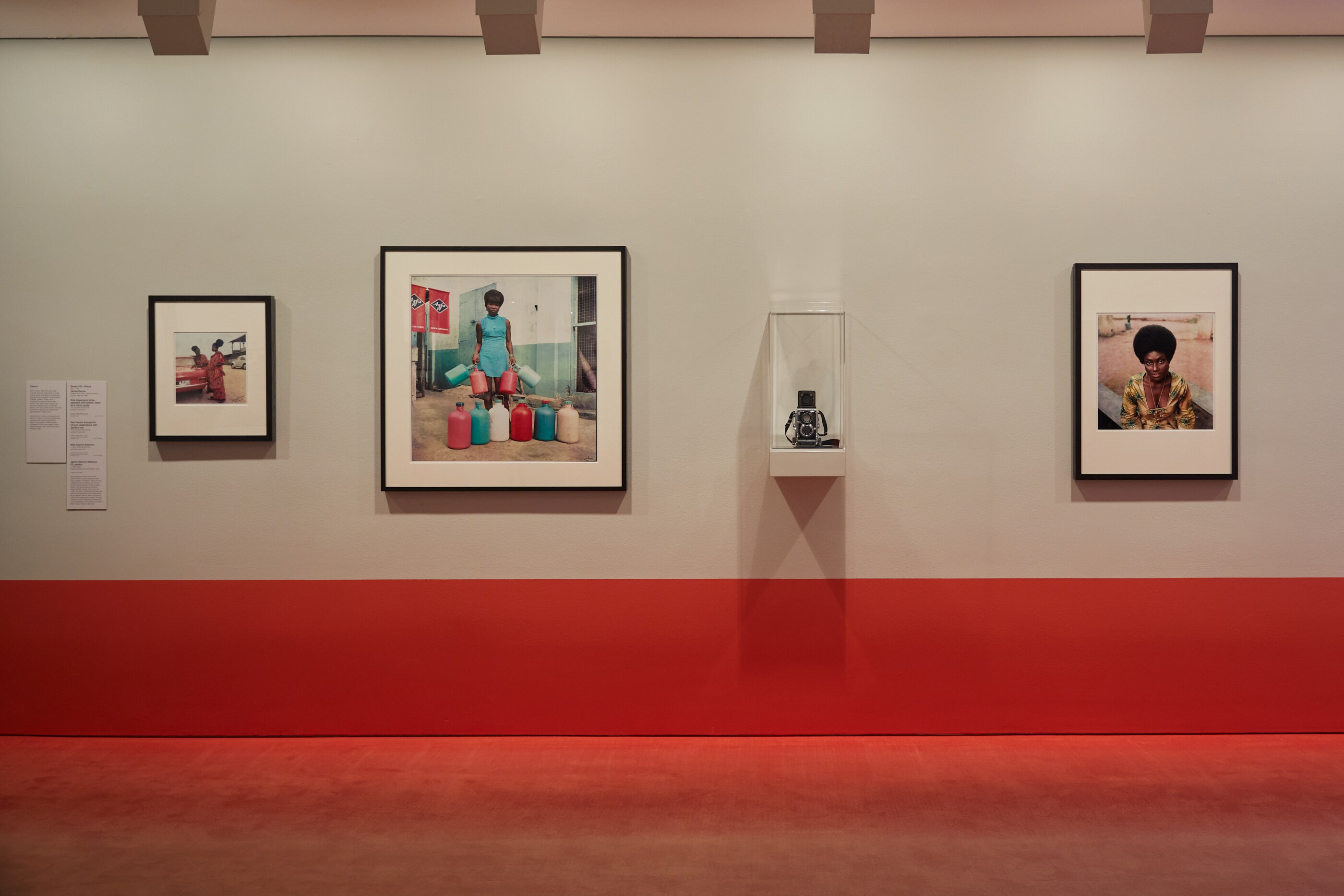 Exhibition wall with three bright colourful photographs of Black women, with a camera in a clear case in the centre. 