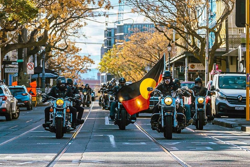 Des motos, dont une portant le drapeau aborigène, passent devant un corbillard noir.