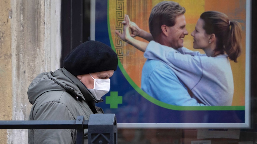 A person wearing a mask walks in front of a large poster depicting a man and a woman in an embrace.