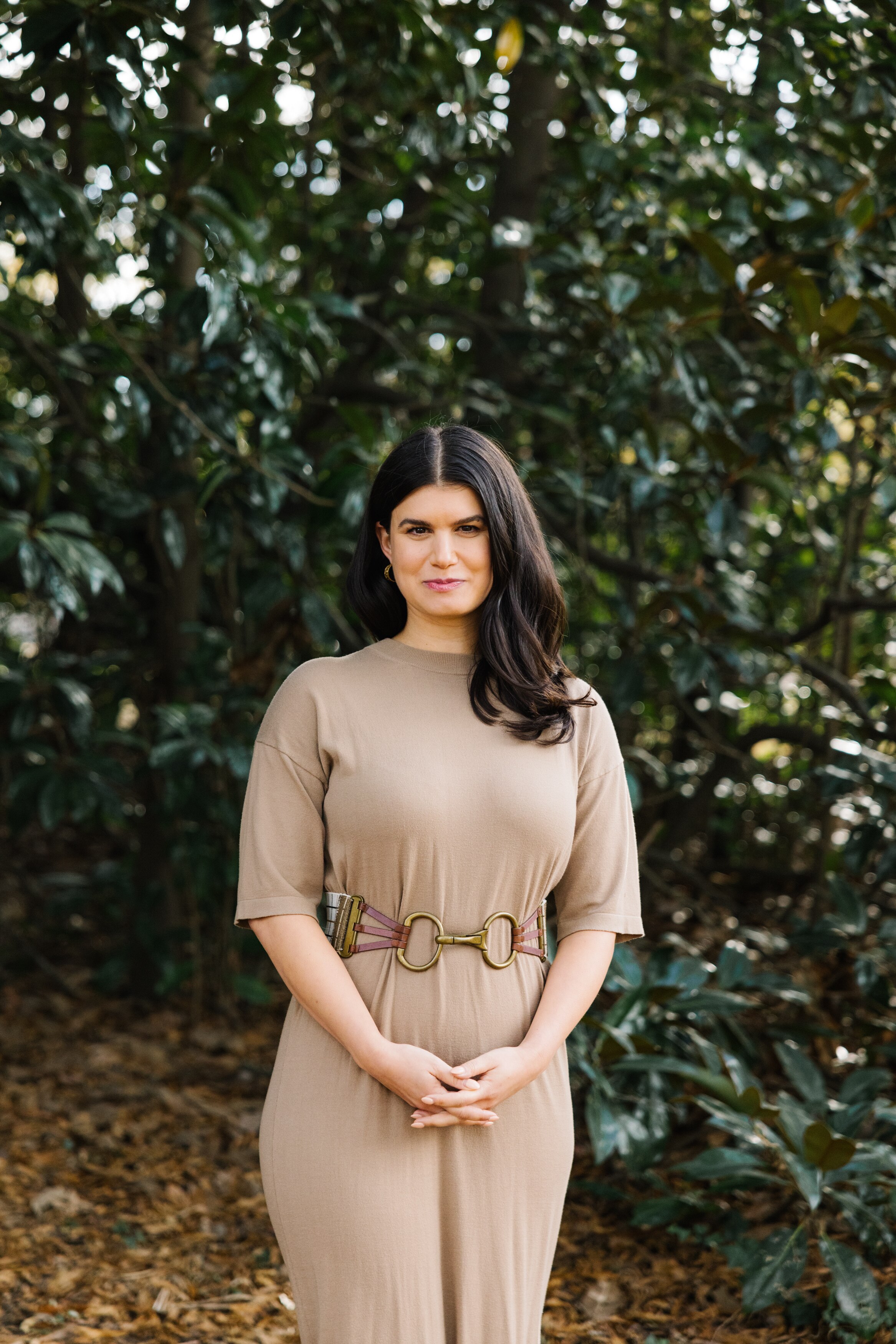 Grief educator Rebecca Feinglos, who has thick brunette hair, poses for a photo in a garden setting wearing a fawn coloured top