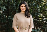 Grief educator Rebecca Feinglos, who has thick brunette hair, poses for a photo in a garden setting wearing a fawn coloured top