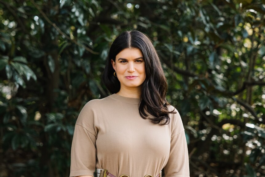 Grief educator Rebecca Feinglos, who has thick brunette hair, poses for a photo in a garden setting wearing a fawn coloured top