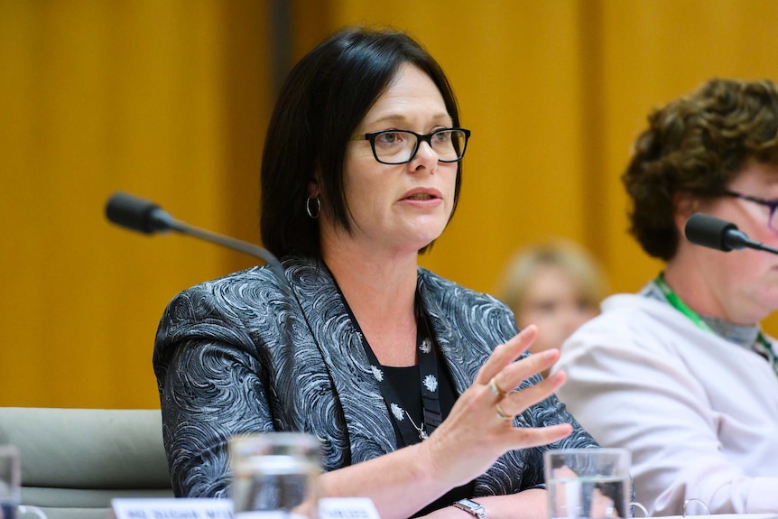 Linda Geddes, wearing glasses and a blazer with a swirly pattern, speaks while sitting at a table at an official hearing.