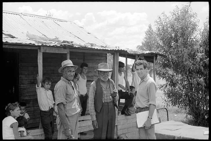 A black and white photo of Charles Perkins standing with two older Aboriginal men. The older men are wearing hats.