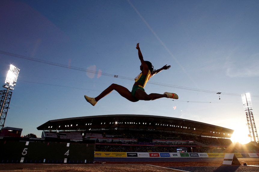 A woman leaps through the air toward a sandpit competing in long jump in front of a grandstand