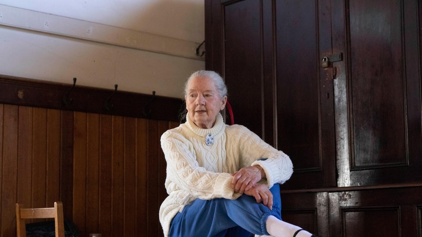 93-year-old Judith Ker sits on a table, in a white knit and ballerina shoes.