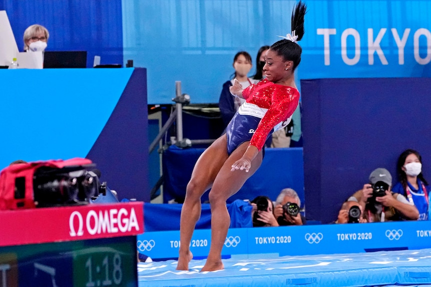 A woman in a leotard lands on a blue mat, her right ankle bent slightly