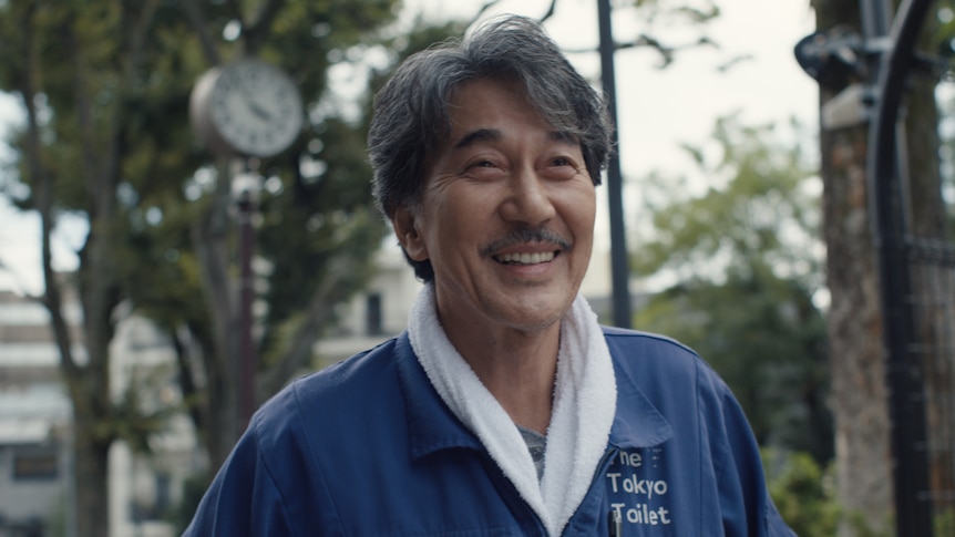 A film still of Kōji Yakusho, a 68-year-old Japanese man, smiling brightly, wearing blue coveralls that read "The Tokyo Toilet".