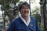 A film still of Kōji Yakusho, a 68-year-old Japanese man, smiling brightly, wearing blue coveralls that read "The Tokyo Toilet".