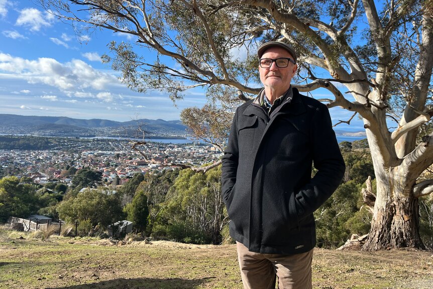 A man in a cap and glasses stands in front of a tree with a view of a city behind