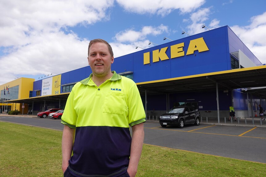 Man with short blonde hair wearing high vis IKEA shirt stands with hands in pockets outside Ikea store.
