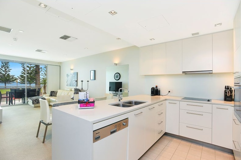 The kitchen and living area of a home. The walls, floor, kitchen and furniture are white, a beach can be seen out the window.