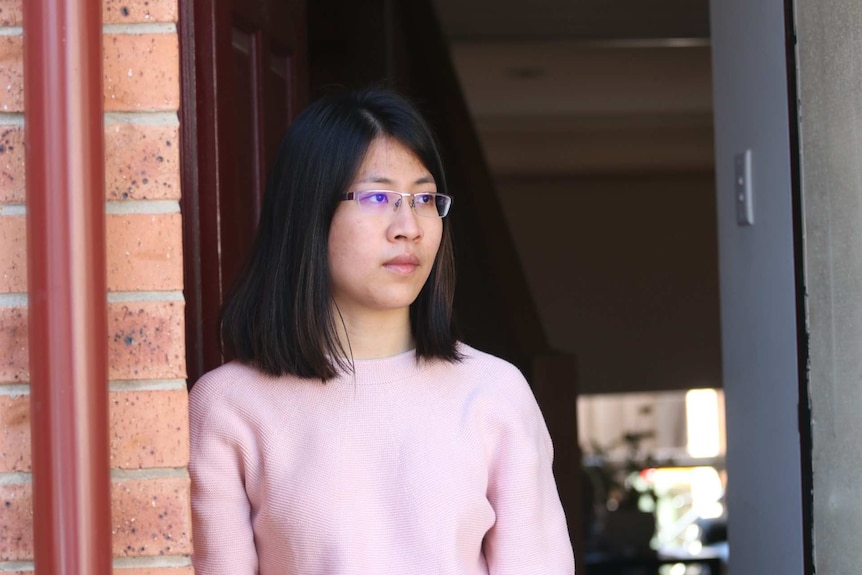 Mien Jeu Chang looks to the side with a serious expression on her face as she stands in front of her door.