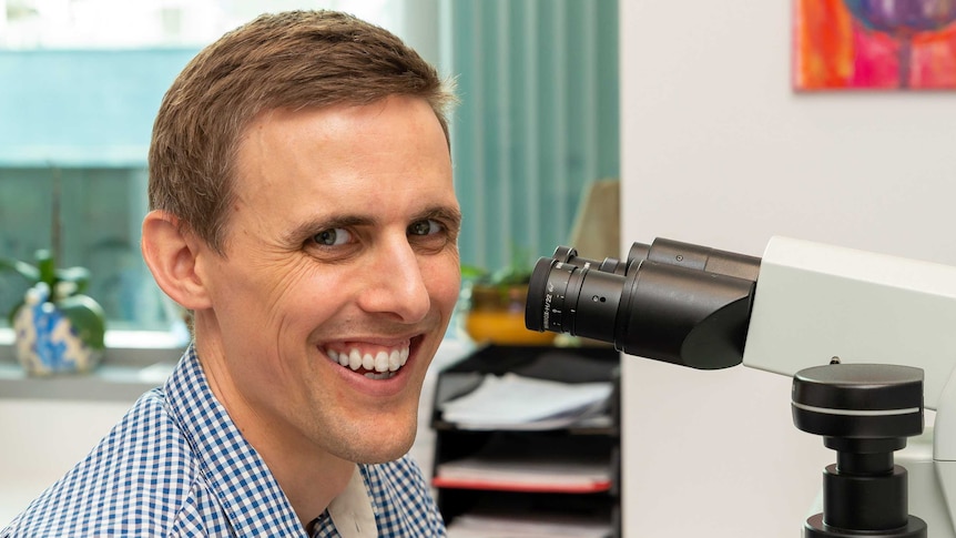 Man in blue and white checked shirt in front of microscope