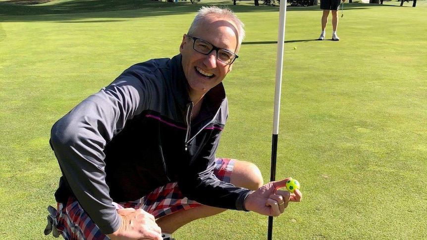 Golfer Jim Grant squats beside a golf hole, smiling and holding a yellow golf ball.