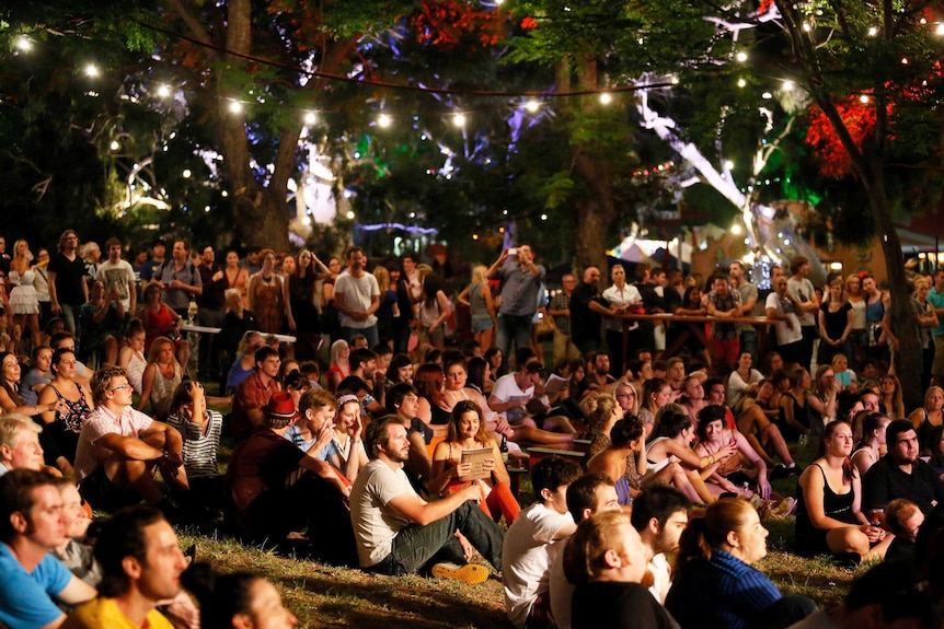 A crowd of people sit on grass under trees lit up with lights.