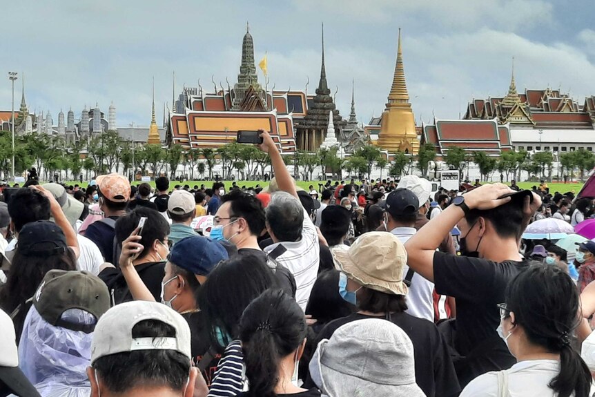 A crowd of people gather taking photos of smartphones with traditional Thai structure in background.