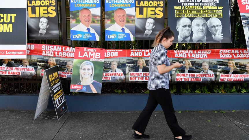A person looking at their phone walks past a fence covered in political posters.