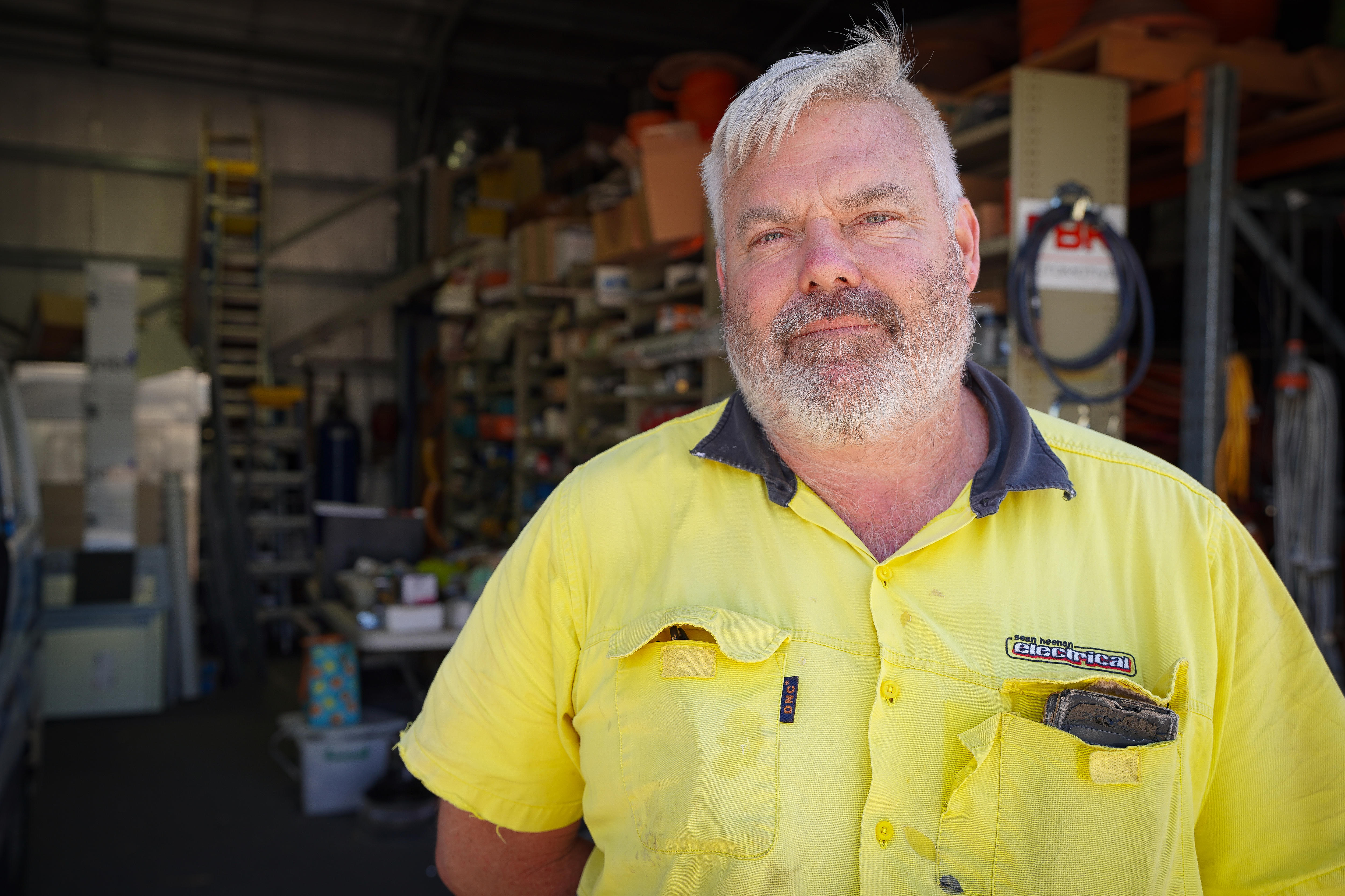 Um homem com barba usa uma camisa de trabalho amarela e uma expressão facial séria. 