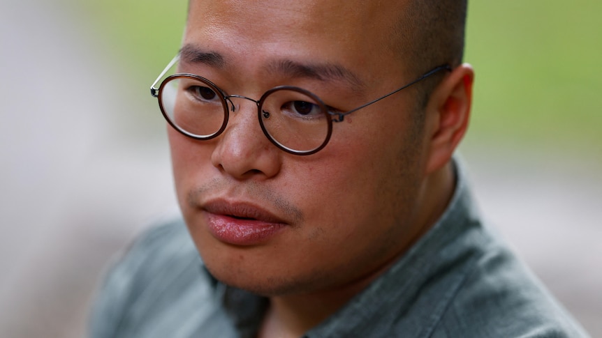 An east-Asian man, receding hair line, round glasses and collard shirt, looks downward.