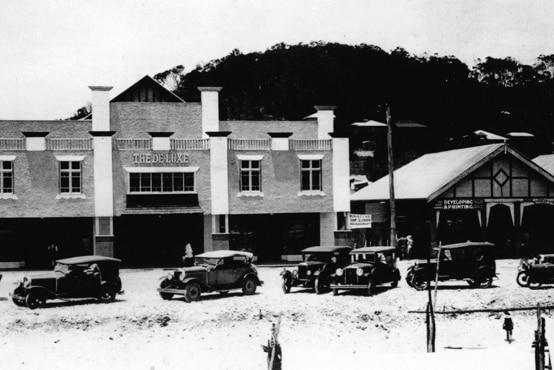 Black and white photo old theatre brick building by a sandy road with old cars.