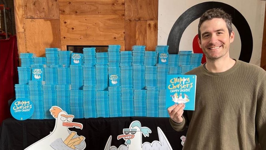 Sam Cotton stands in front of a pile of his new blue books, witha  seagull illustration