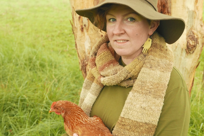 Uma mulher séria usando um chapéu verde segurando uma galinha fica contra uma árvore na grama verde.