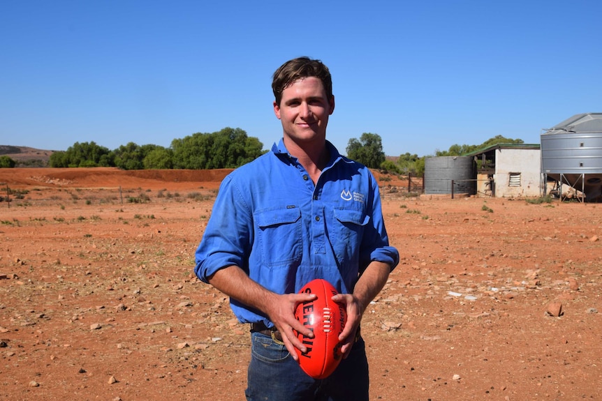 A man in a blue rolled up work shirt holds a red AFL ball. behind him is red dirt, shubbery and silcer water tanks to the left.