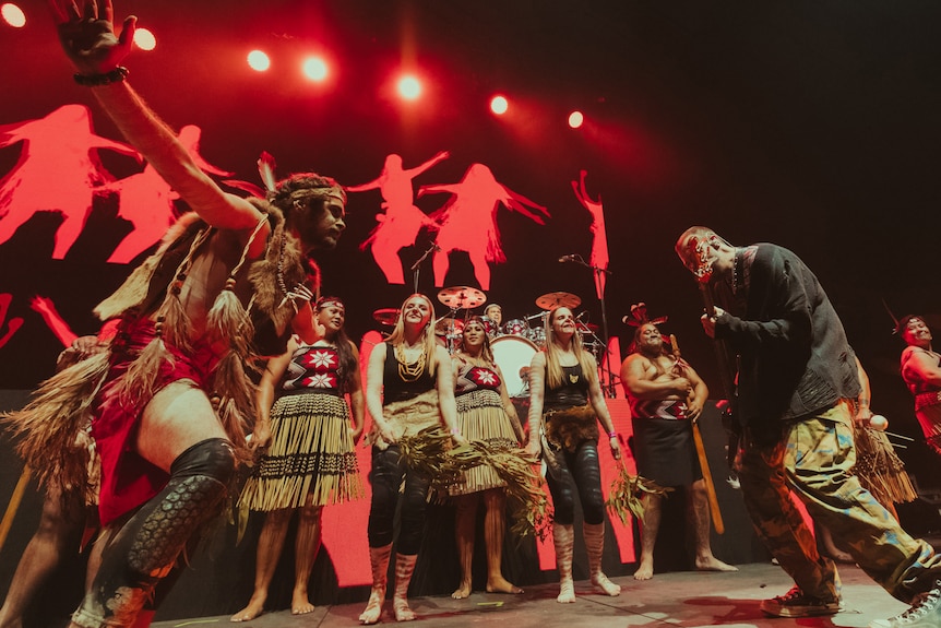 Le bassiste SIX60 Chris Mac avec des danseurs de Nunukul Yuggera sur scène éclairée en rouge portant des vêtements traditionnels maoris et aborigènes