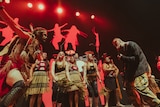 SIX60 bass player Chris Mac with Nunukul Yuggera dancers on red lit stage wearing traditional maori and Aboriginal dress