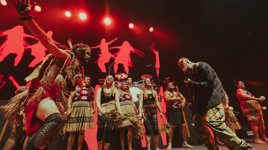 SIX60 bass player Chris Mac with Nunukul Yuggera dancers on red lit stage wearing traditional maori and Aboriginal dress