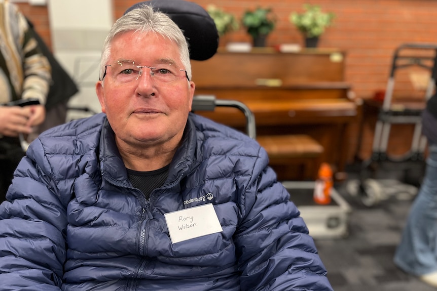 Man wearing glasses and a puffer jacket sits in a wheelchair, looking pleasantly at the camera