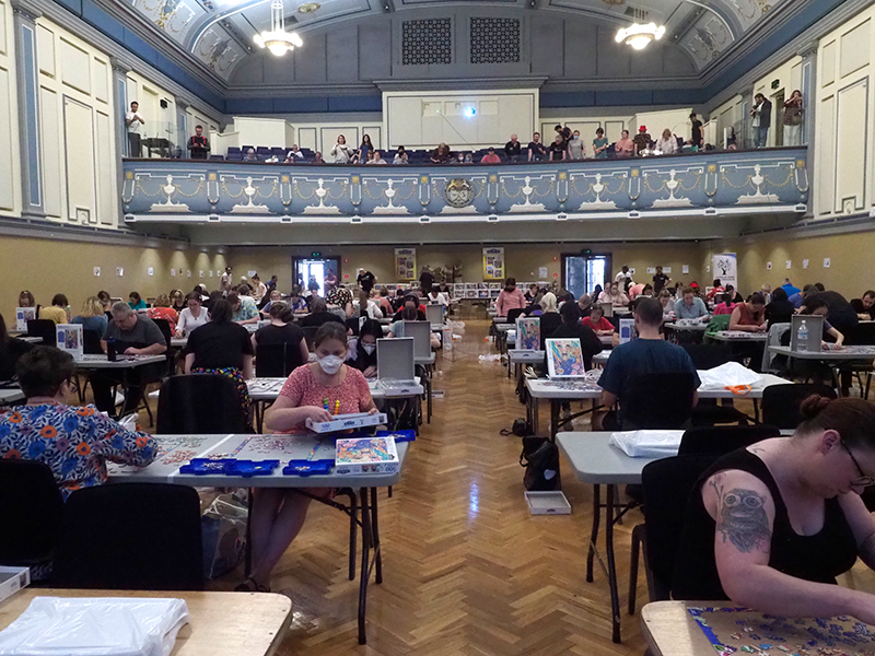 A hall full of people sitting at tables doing jigsaw puzzles under competition deadline