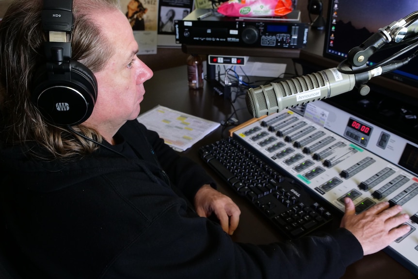 A Man sits at radio desk, headphones on, hand on fader, microphone suspended on arm.