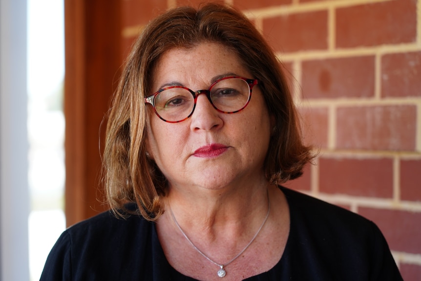 A woman with shoulder length brown hair and glasses stands in front of a brick wall