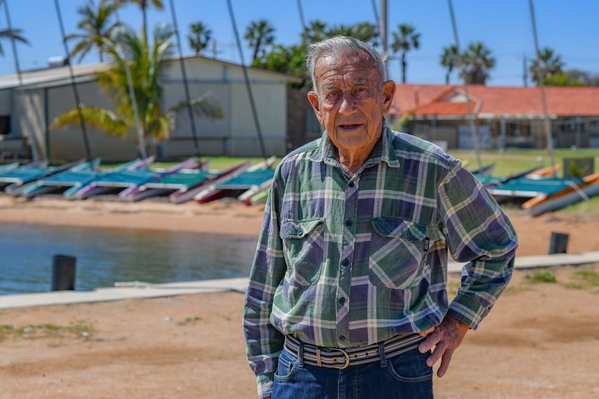 Elderly man stands facing camera in checkered shirt. Palm trees and sailing boats in background