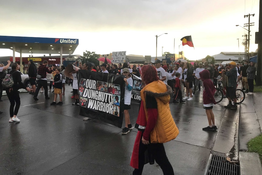A group of protesters, waving Aboriginal flags and carrying placards, walk past a service station on a rain-slicked street.