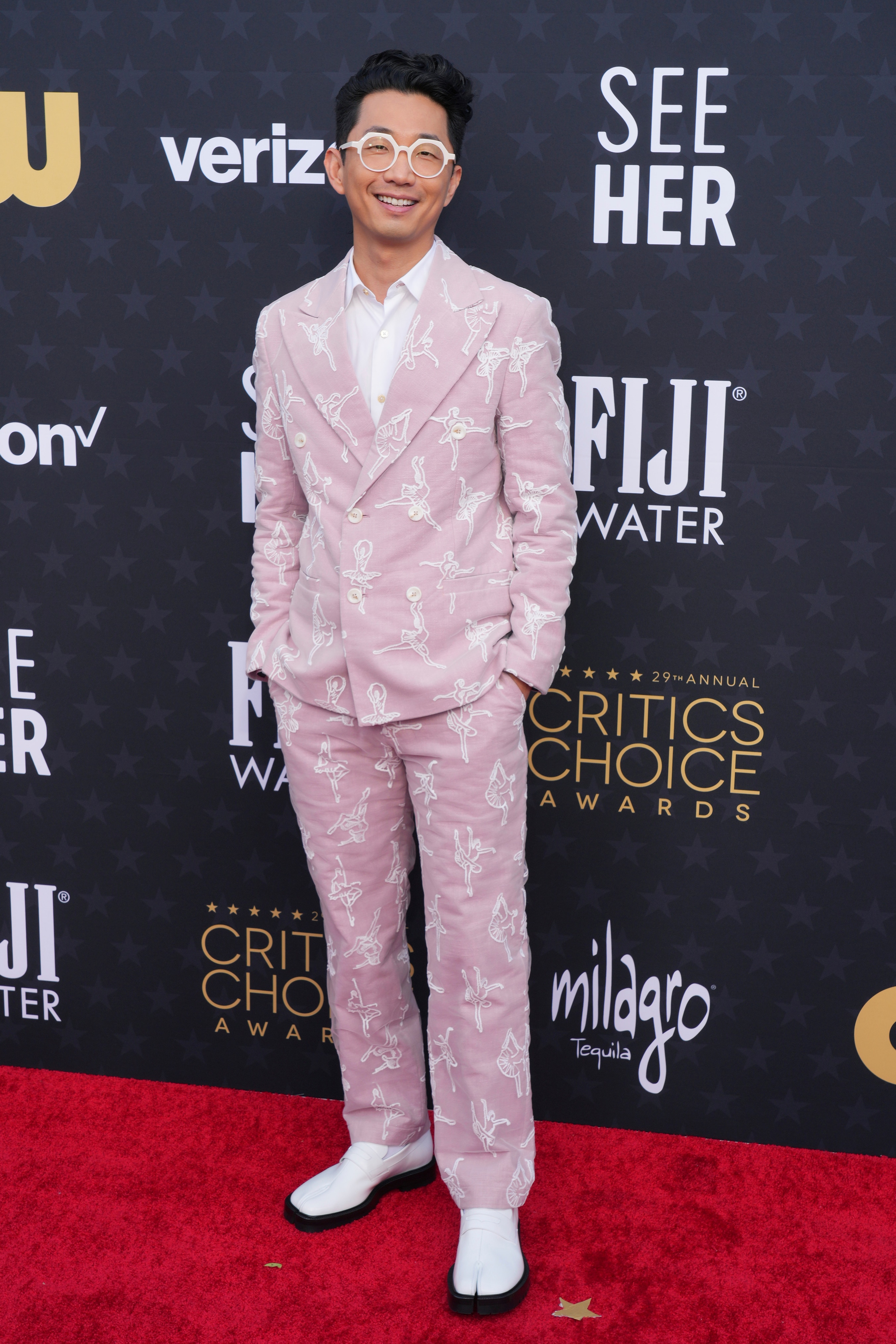 Lee Sung Jin wearing a pastel pink suit with white ballerinas printed on the fabric, which glasses and white shoes. 