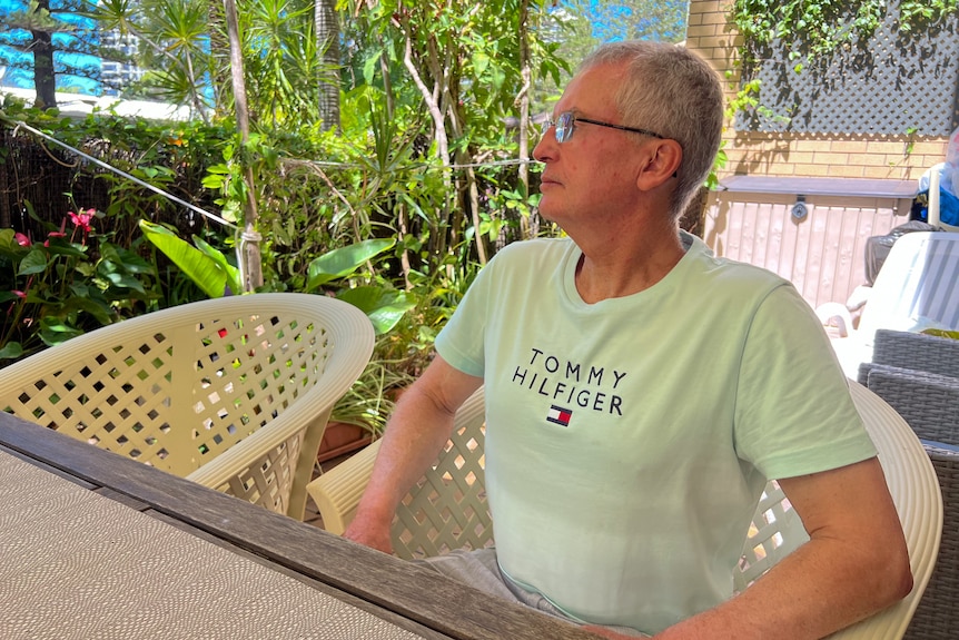 A man in a mint green t-shirt sits at an outdoor table near a garden.