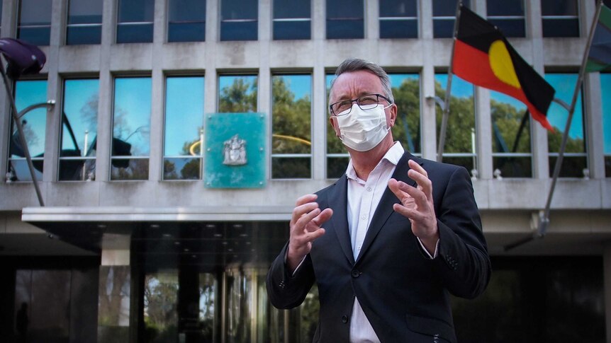 Mental Health Minister Martin Foley stands in front of a building with an Indigenous flag waving. He wears a face mask and suit.