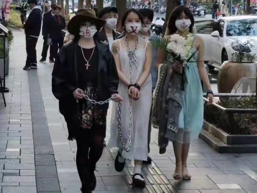Three women walk down a street with a chain attached to one in a white dress. They all wear masks