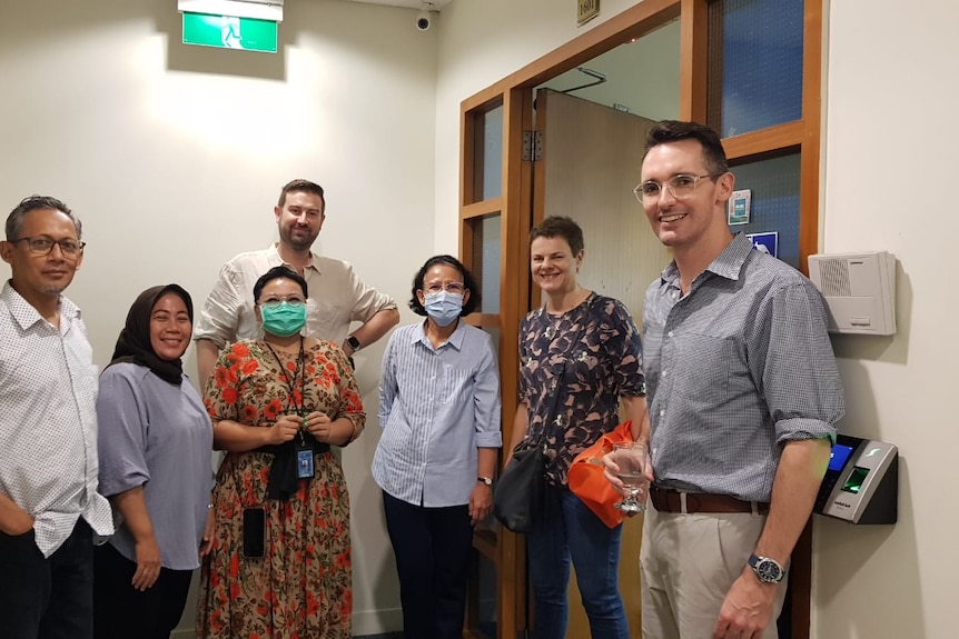 Three men and four women, some wearing masks, standing in an office doorway posing for a photo.