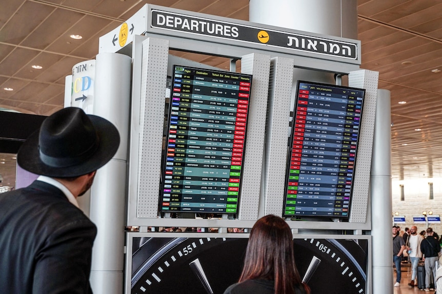 People stand and watch the flight board which has many delayed flights at the airport.