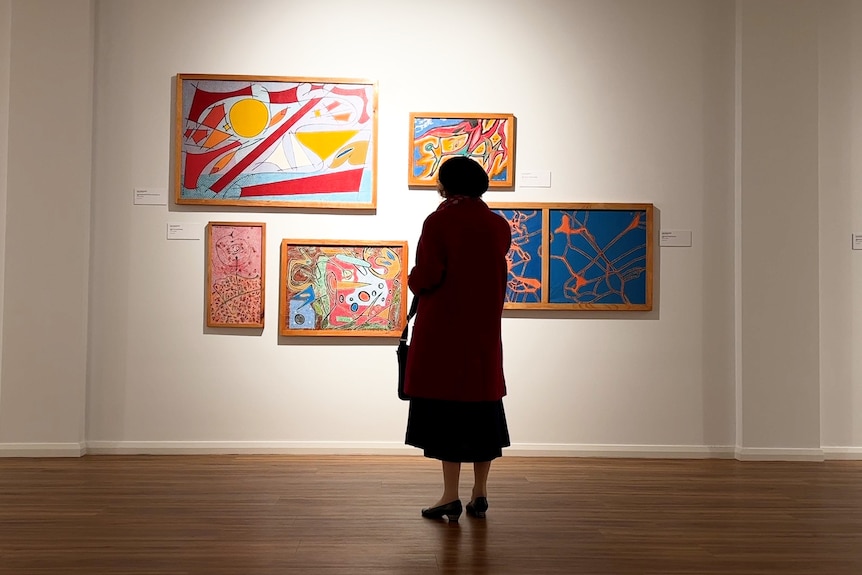 A well dressed older woman stands silhouetted in front of a wall of modern artworks