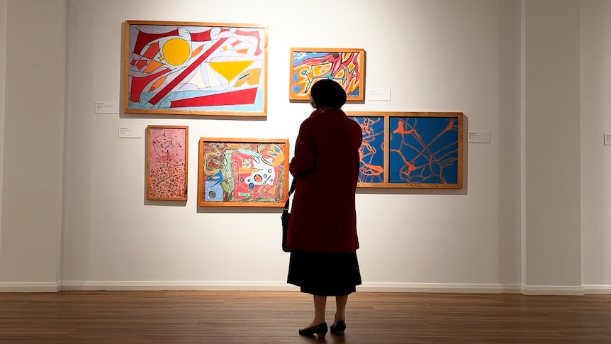 A well dressed older woman stands silhouetted in front of a wall of modern artworks
