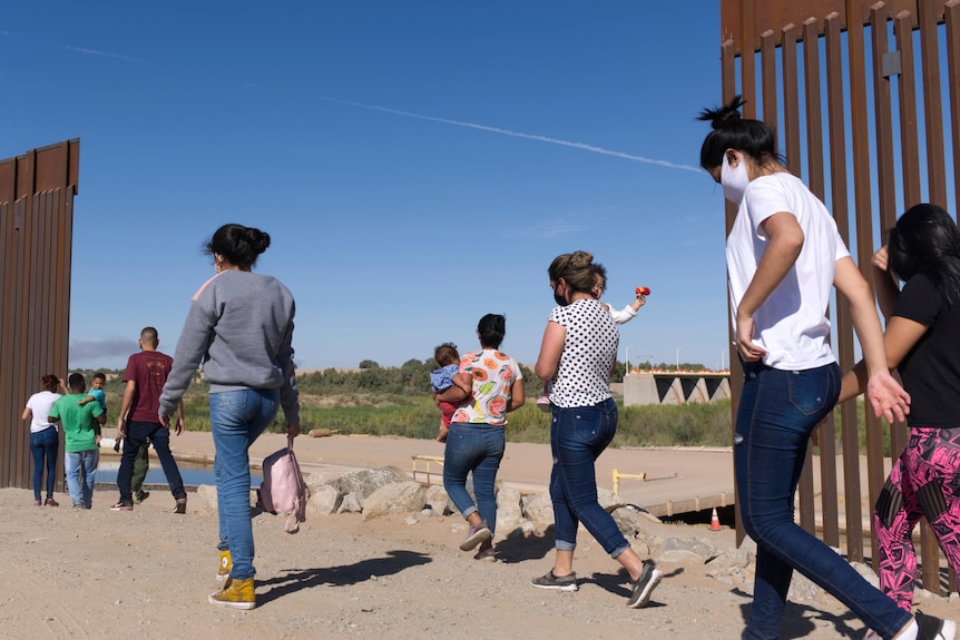 Women and children wearing casual clothing walk through a gap in a red metal fence in the Arizona desert.