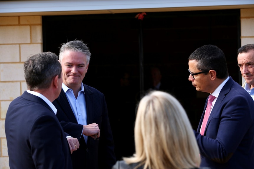 Mathias Cormann stands wearing an unbuttoned shirt and jacket facing the camera next to Mark McGowan and Ben Wyatt.