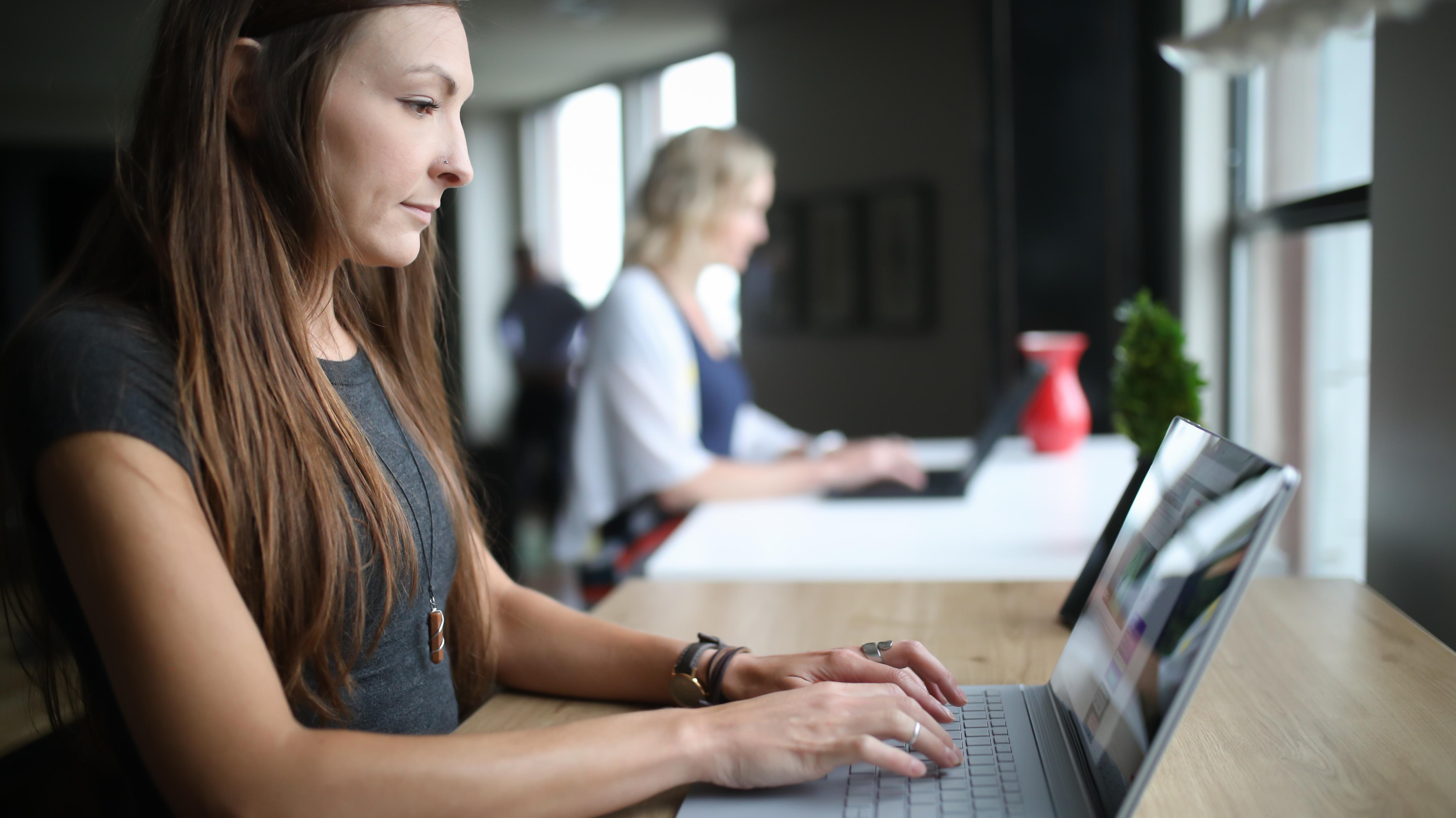 A woman with long brown hairis at a standup desk in focus. Another woman on another computer with blonde hair can be seen