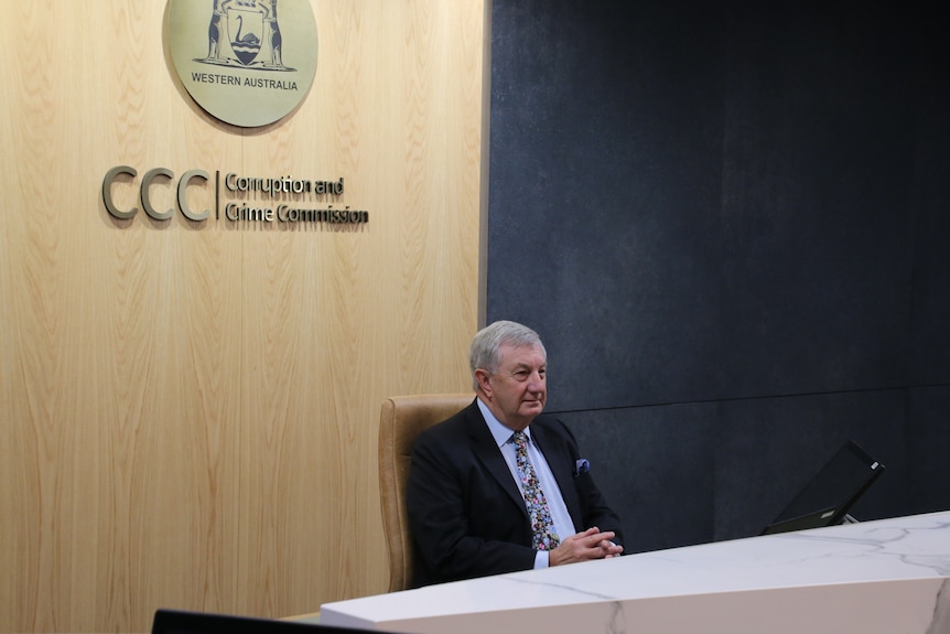 A man sits in a hearing room with the letters CCC and Corruption and Crime Commission signage.